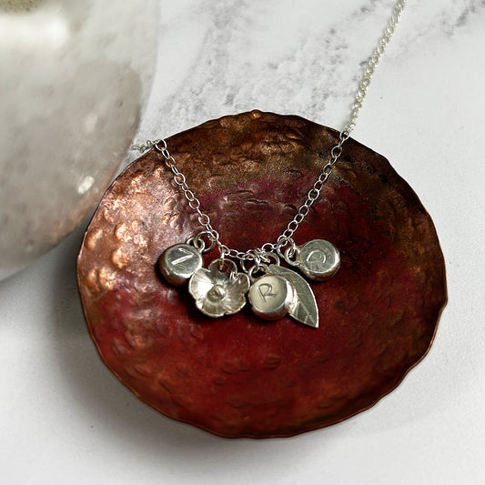 An image of a handmade silver necklace showing a flower, a leaf, and three silver pebbles with the letters Z, R, and D stamped on them.