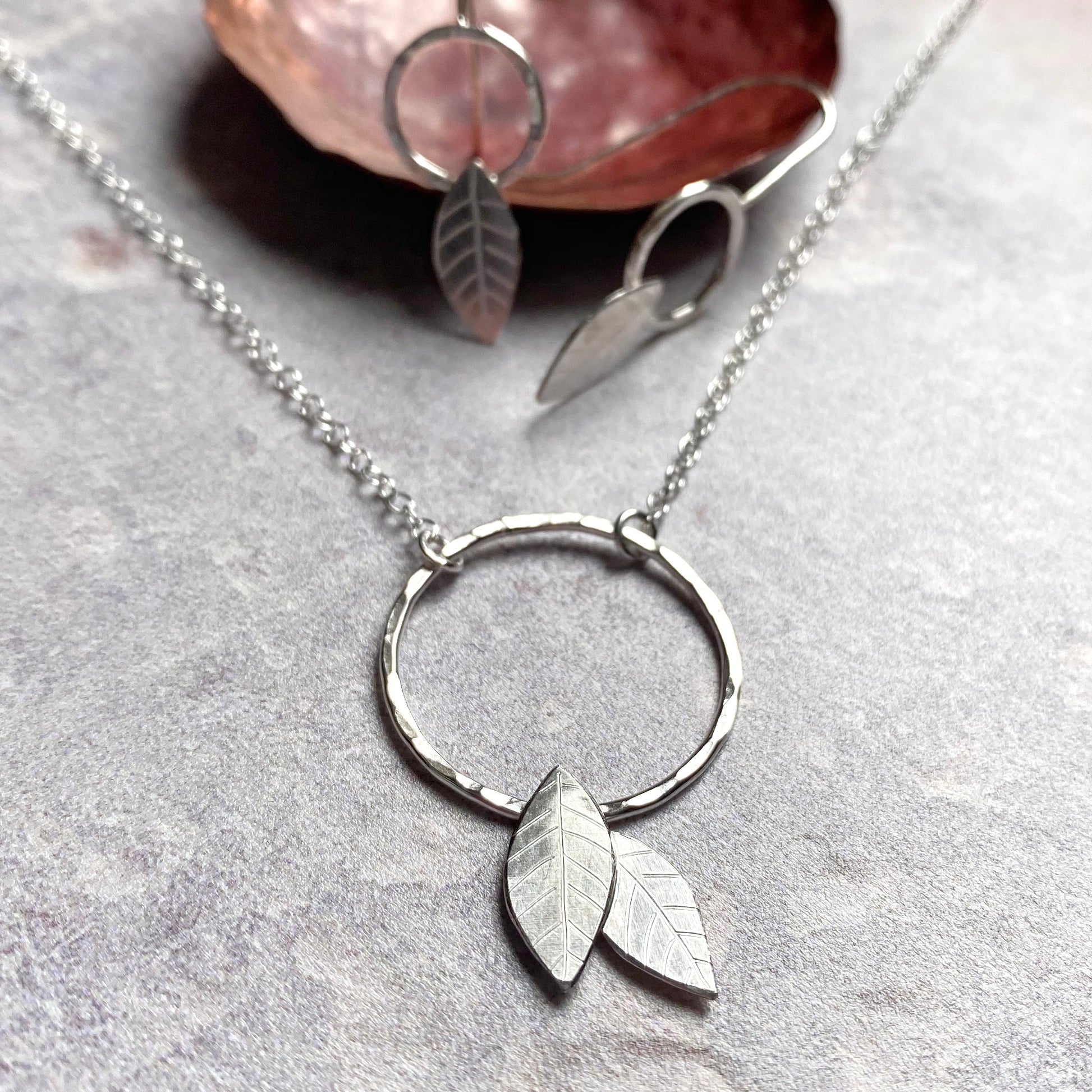 A handmade silver pendant of an open silver circle with two 2D silver leaves attached if falling off of the bottom, with a complimentary pair of earrings, by zoe ruth designs