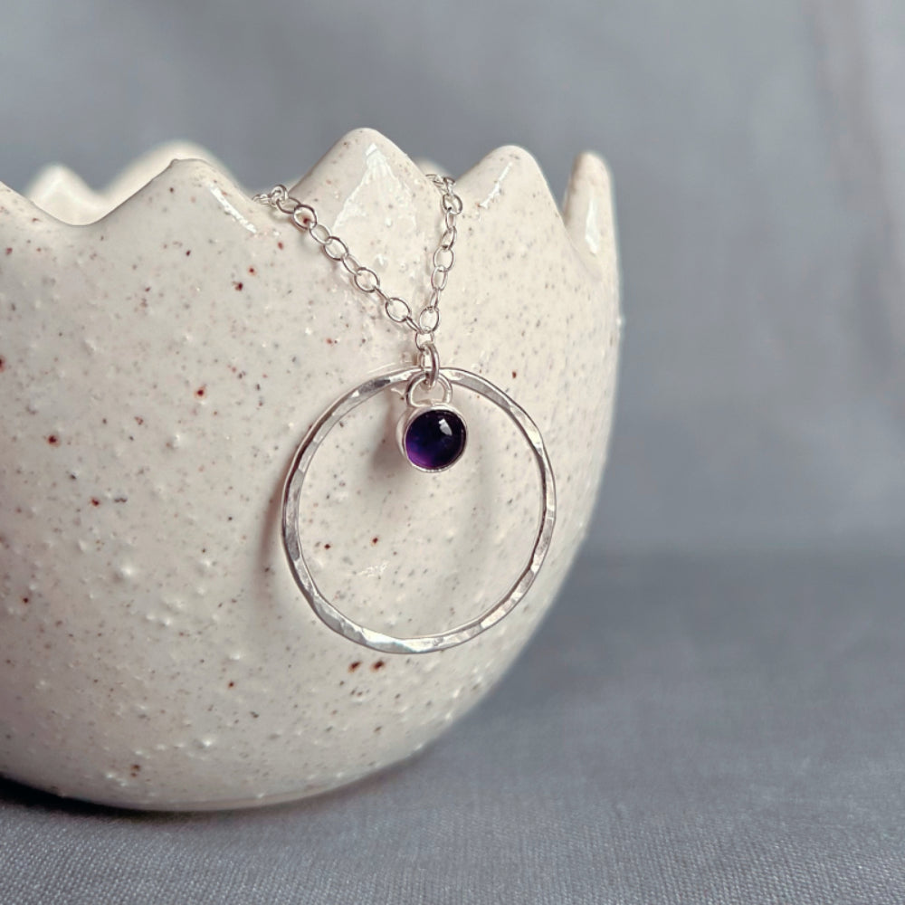 A 5mm deep purple Amethyst stone set in silver hangs loosely at the top of a larger slim and hammered circle frame as the pendant of this necklace by zoe ruth designs.