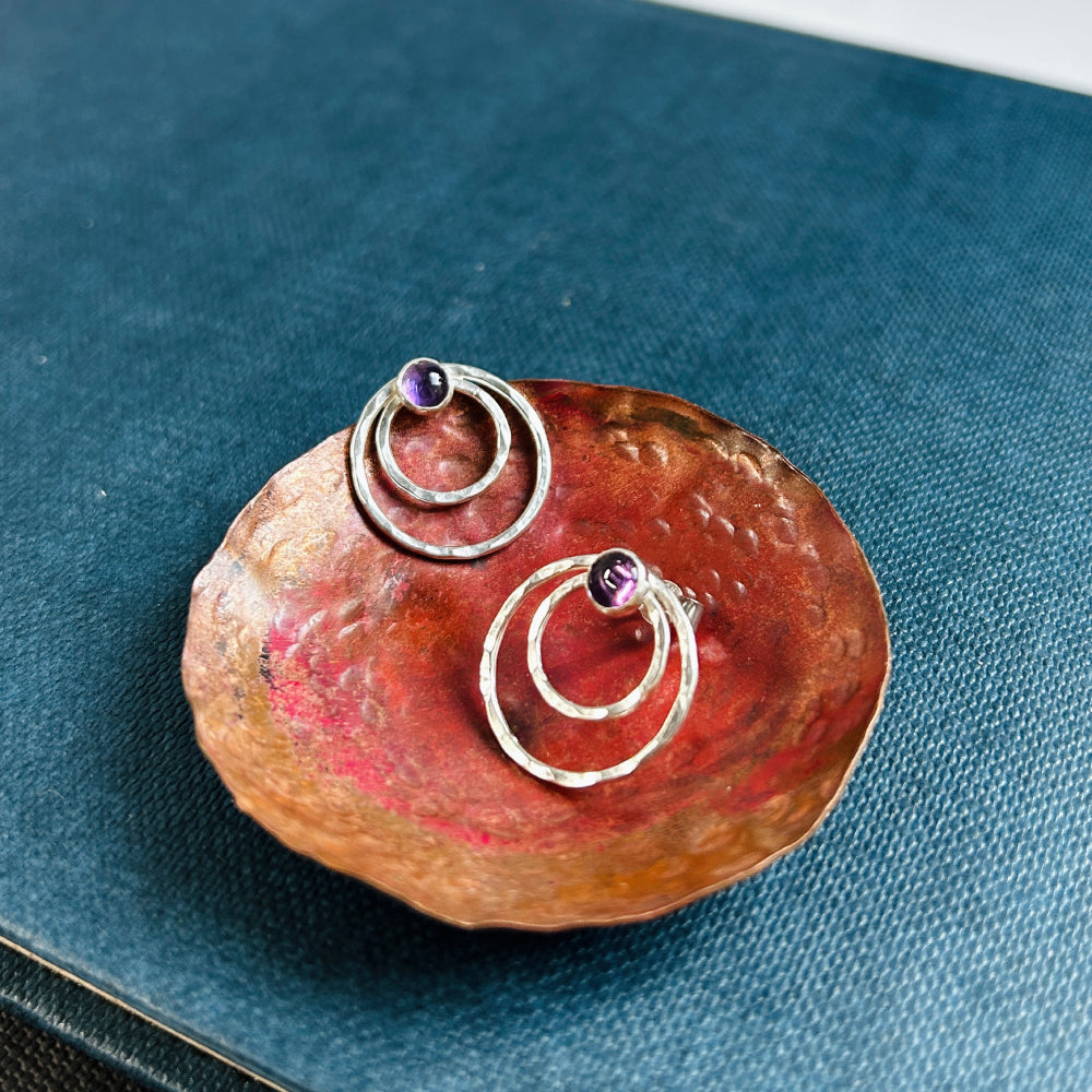 Two small concentric circles handmade in hammered silver make each of these stud earrings, with a small amethyst cabochon at the top of each earring.  Displayed sat in a copper dish