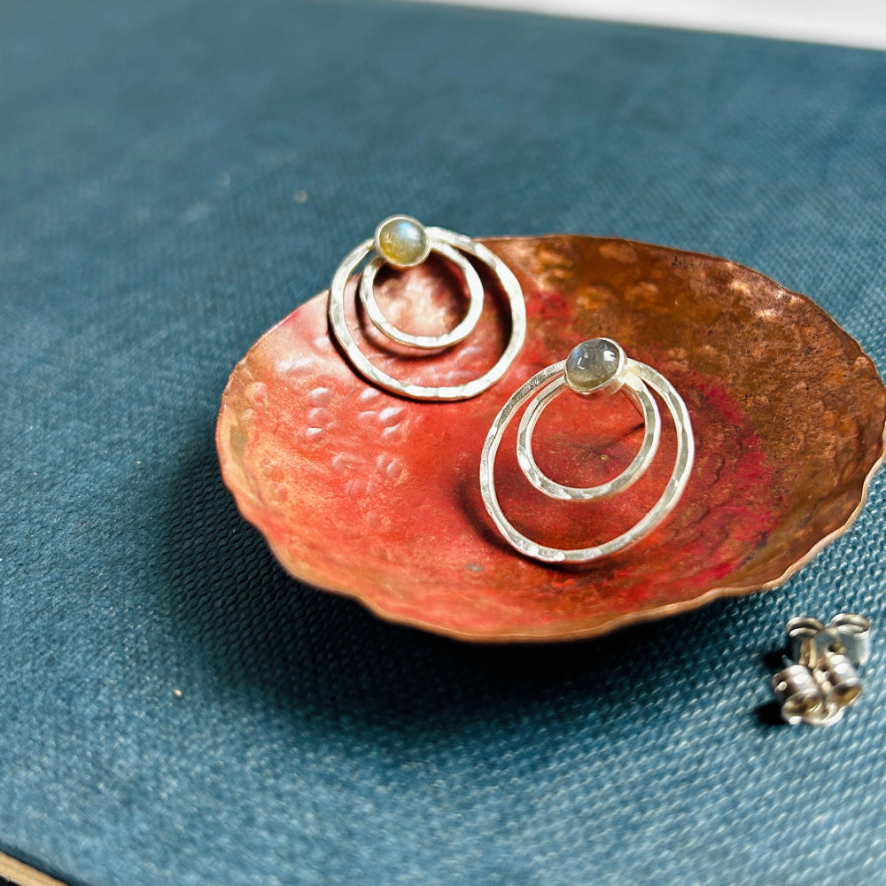 Two small concentric circles handmade in hammered silver make each of these stud earrings, with a small labradorite cabochon at the top of each earring. Displayed sat in a copper dish