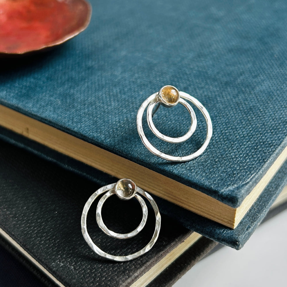 Two small concentric circles handmade in hammered silver make each of these stud earrings, with a small smokey quartz cabochon at the top of each earring.  Displayed sat at different angles on a pile of old books