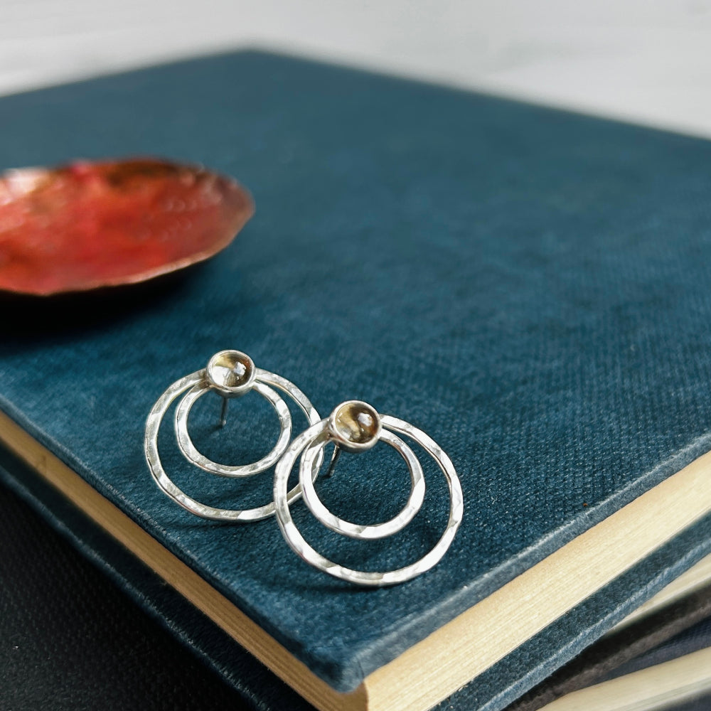 Two small concentric circles handmade in hammered silver make each of these stud earrings, with a small smokey quartz cabochon at the top of each earring. Sat on an old blue book with a copper dish in the background