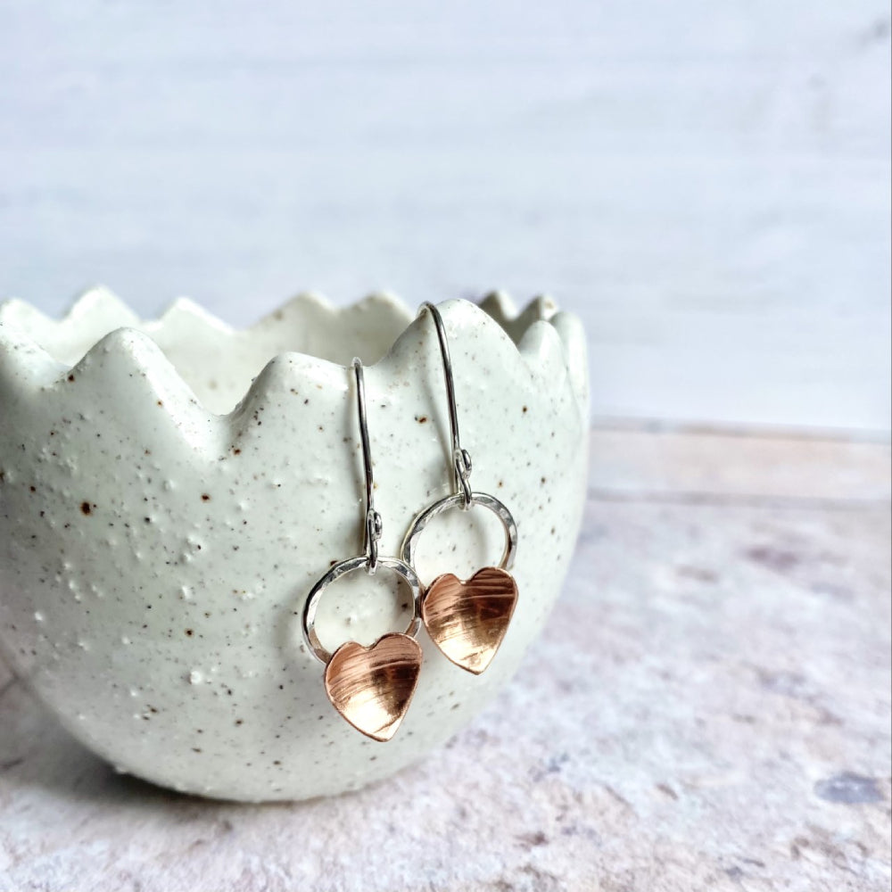 A pair of handmade copper heart and silver circle hook earrings hung over a cream ceramic pot, by zoe ruth designs.