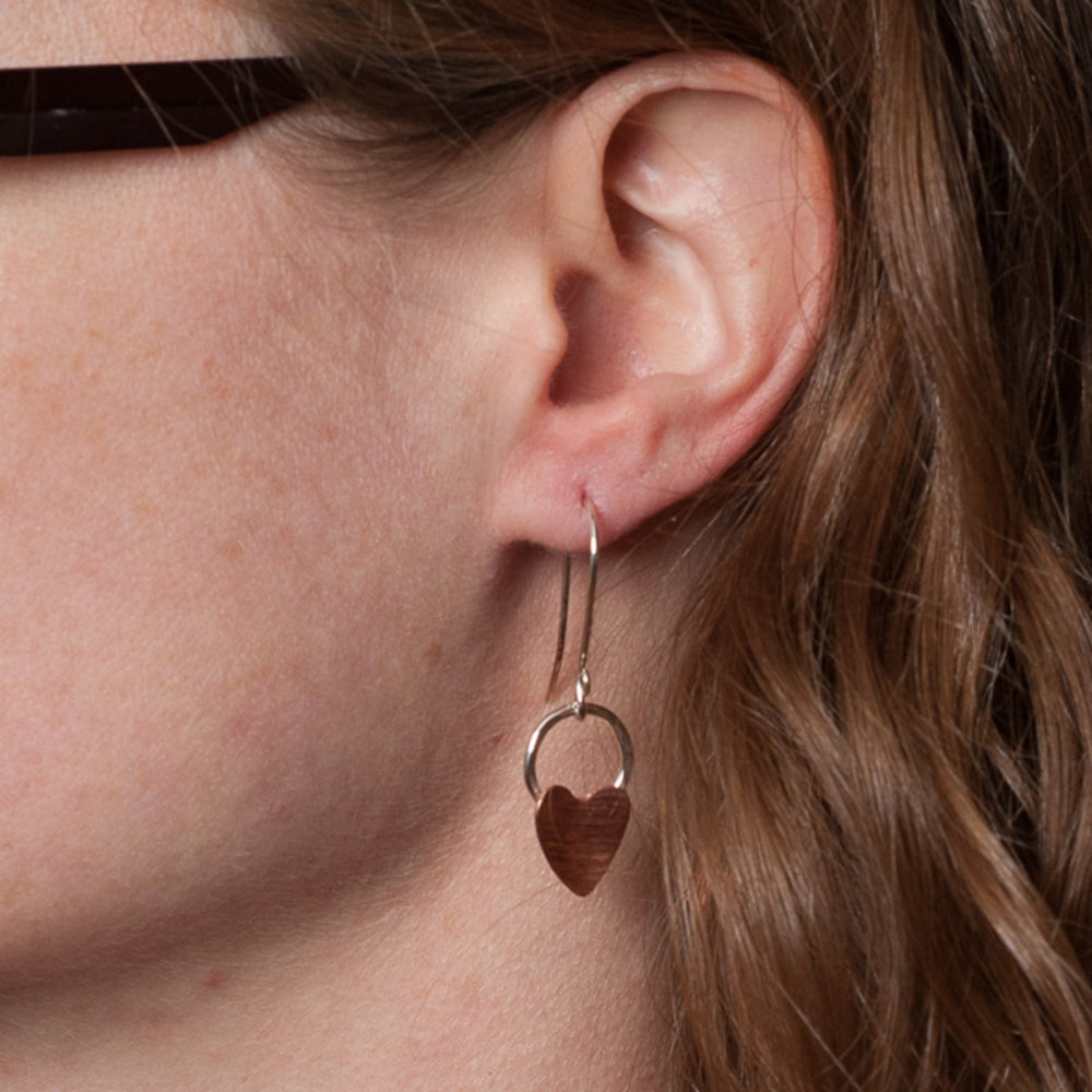 A person wearing a hook style earring, it has a long hook drop with a copper heart attached to a silver circle dangling from it.