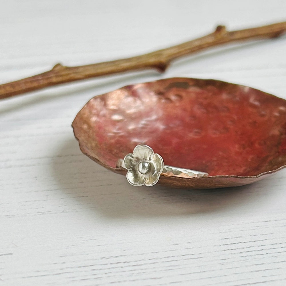 A five petaled silver flower sits on a hammered silver slim ring, balanced on the edge of a copper dish, by zoe ruth designs