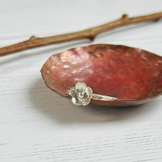A five petaled silver flower sits on a hammered silver slim ring, balanced on the edge of a copper dish, by zoe ruth designs