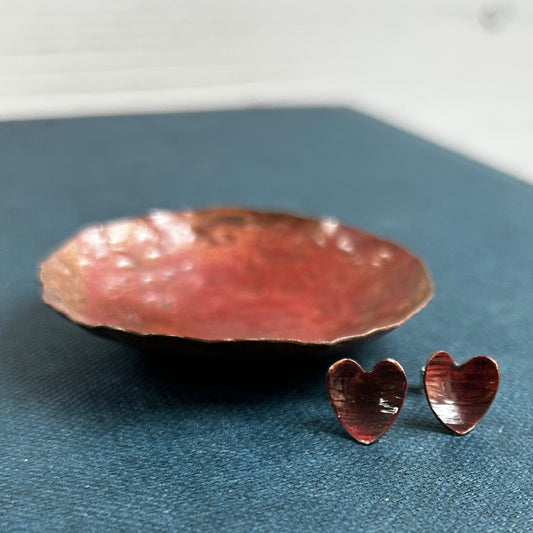 Dark reddish brown copper heart shaped stud earrings, sat on a blue book and next to a copper dish, by zoe ruth designs