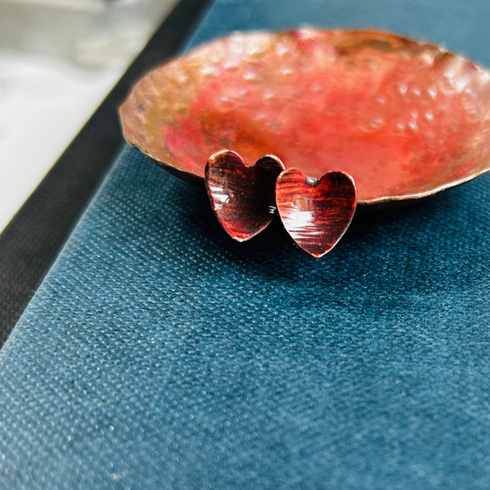 Dark reddish brown copper heart shaped stud earrings, sat on a blue book and next to a copper dish, by zoe ruth designs