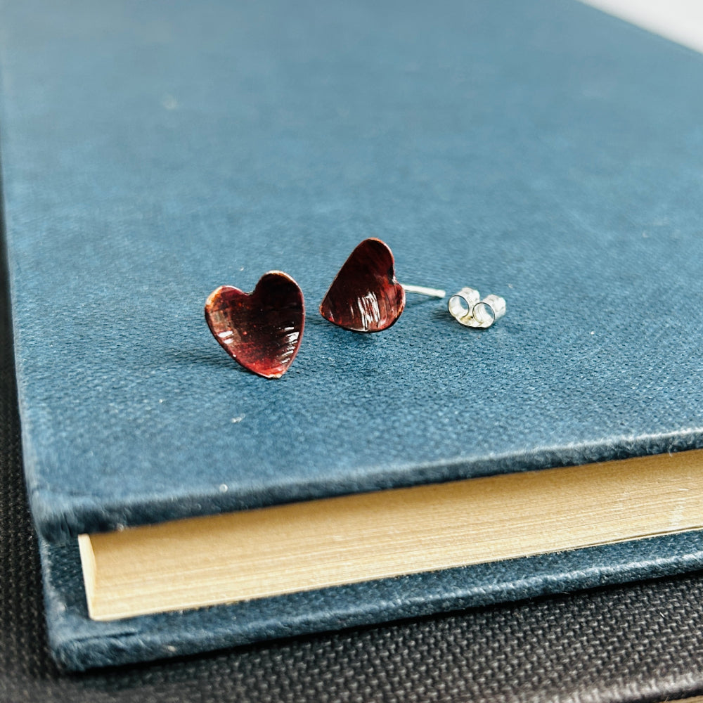 Dark reddish brown copper heart shaped stud earrings, sat on a blue book, by zoe ruth designs