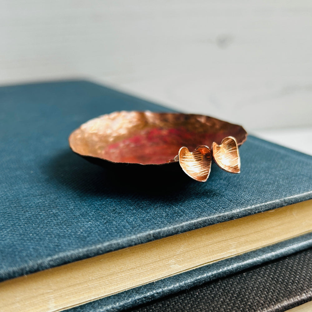 Heart shaped hammered with a lines texture copper stud earrings, sat on a copper dish on a blue book, by zoe ruth designs