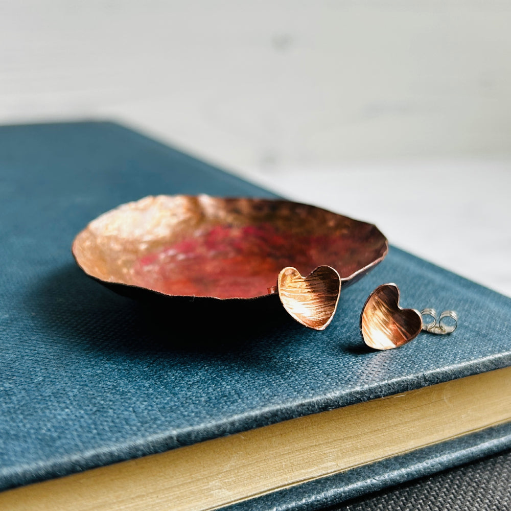 Heart shaped hammered with a lines texture copper stud earrings, sat on a copper dish on a blue book, by zoe ruth designs