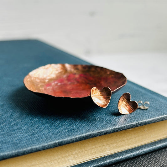 Heart shaped hammered with a lines texture copper stud earrings, sat on a copper dish on a blue book, by zoe ruth designs