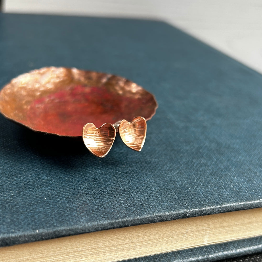 Heart shaped hammered with a lines texture copper stud earrings, sat on a copper dish on a blue book, by zoe ruth designs