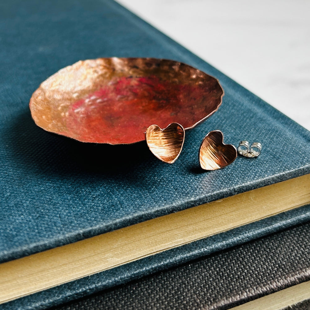 Heart shaped hammered with a lines texture copper stud earrings, sat on a copper dish on a blue book, by zoe ruth designs