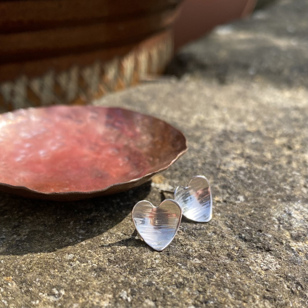 A pair of heart shaped silver earrings with a linear hammered pattern on the surface sit oouitside on a concrete wall top next to a copper dish, by zoe ruth designs