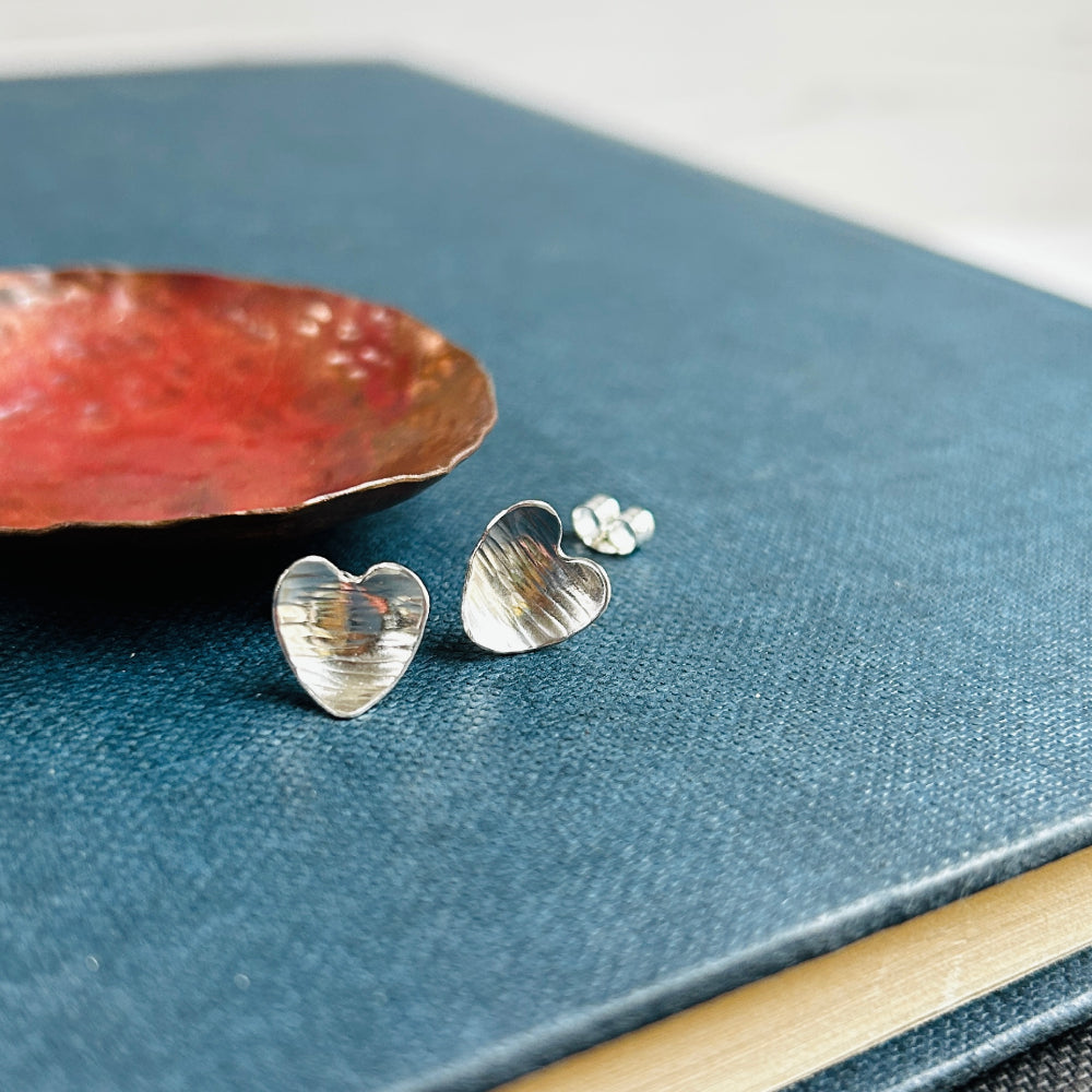 A pair of heart shaped silver earrings with a linear hammered pattern on the surface sit on a blue book next to a copper dish, by zoe ruth designs