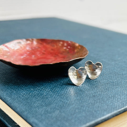 A pair of heart shaped silver earrings with a linear hammered pattern on the surface sit on a blue book next to a copper dish, by zoe ruth designs