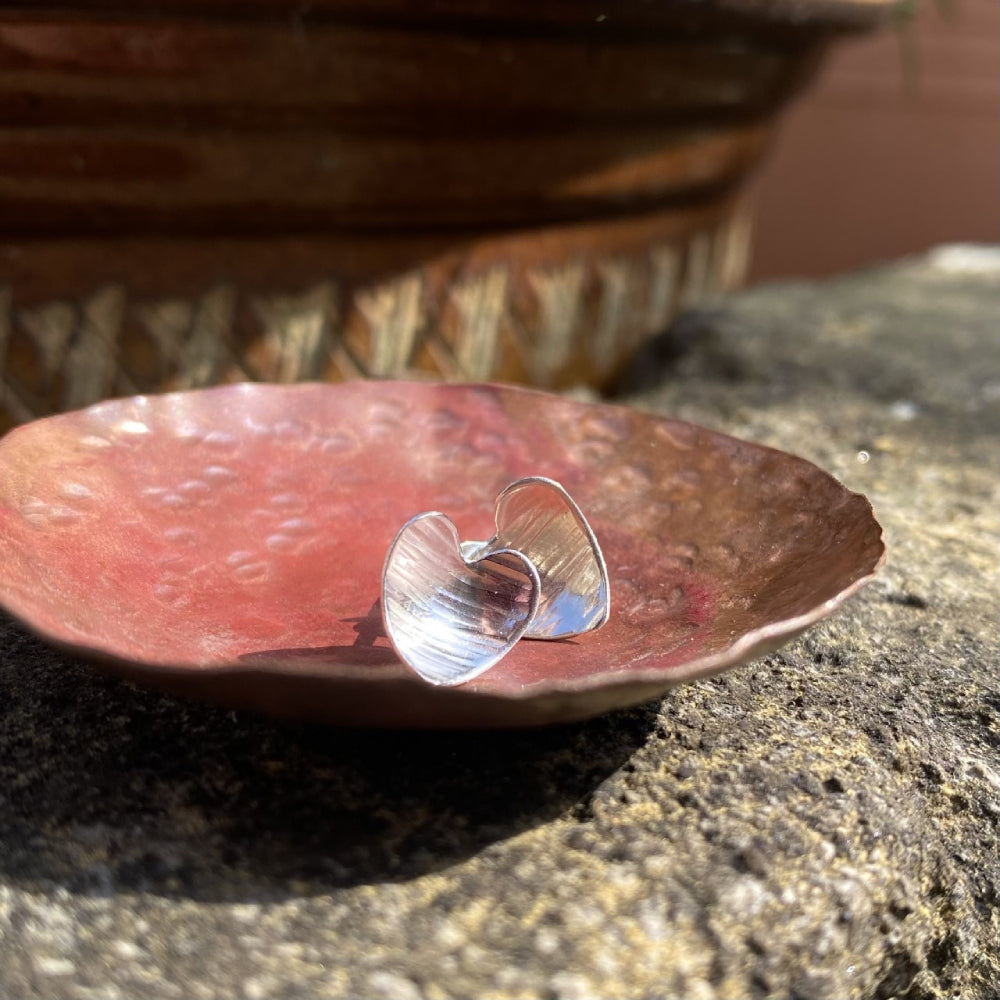 A pair of heart shaped silver earrings with a linear hammered pattern on the surface sit in a copper dish in the sunshine, by zoe ruth designs
