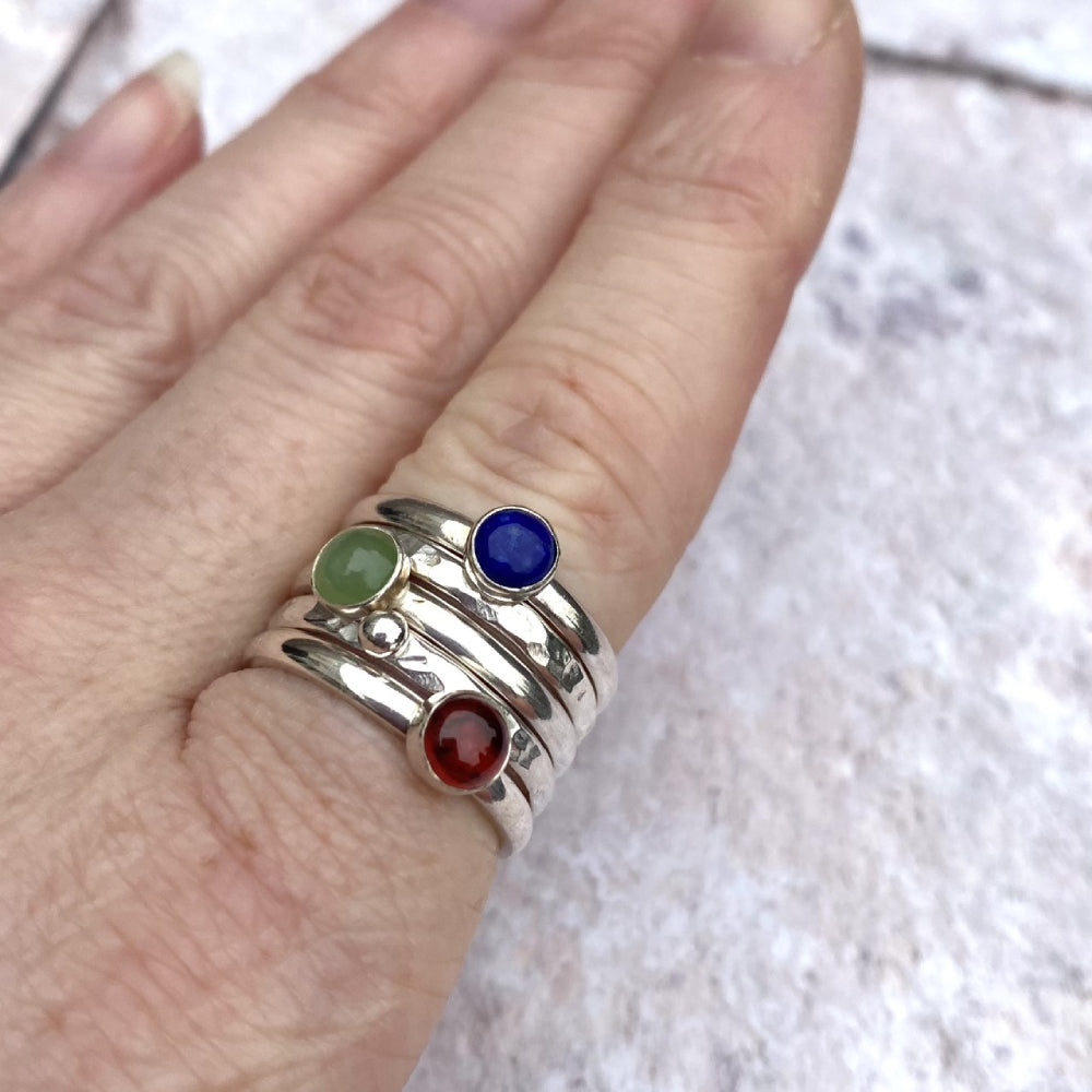 A stack of five silver rings, two textured and one of those with a silver pebble, three smooth with stones set on top, a green Jade, a blue lapis lazuli, and a red garnet, by zoe ruth designs