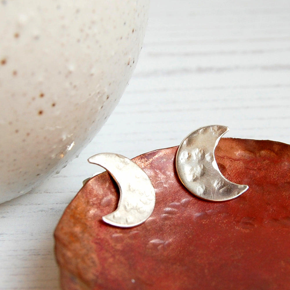 A pair of hand sawn and textured sterling silver crescent moon shaped stud earrings, perched on the edge of a copper dish, by zoe ruth designs