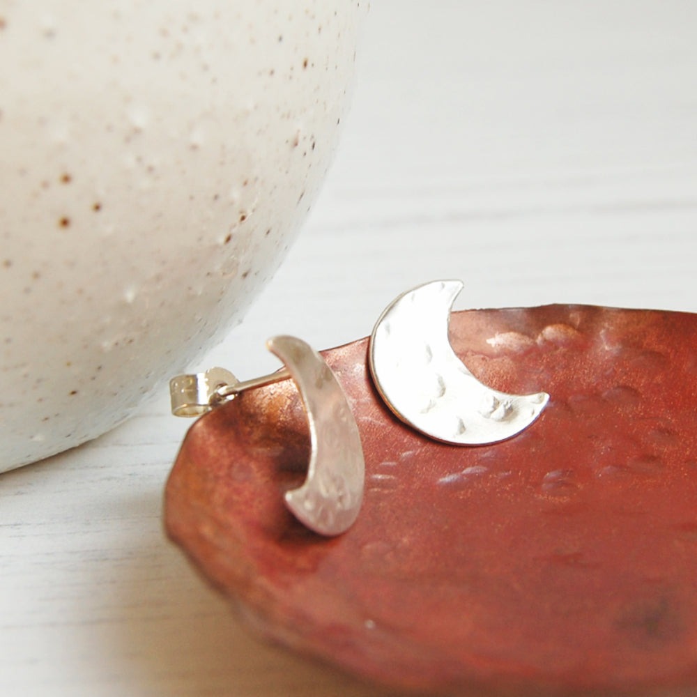 A pair of hand sawn and textured sterling silver crescent moon shaped stud earrings, sat in a copper dish, by zoe ruth designs