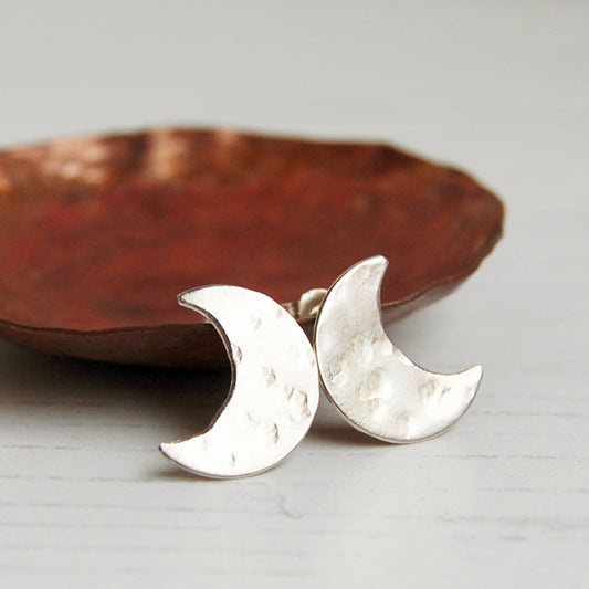 A pair of hand sawn and textured sterling silver crescent moon shaped stud earrings, infront of a copper dish, by zoe ruth designs
