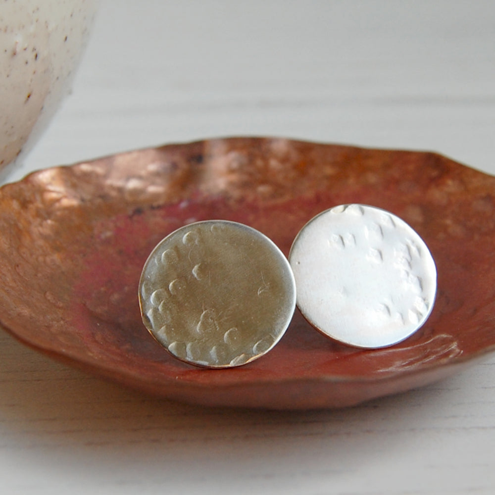 A pair of handmade full moon silver studs, flat silver disks with a lightly hammered surface, sat in a copper dish, by zoe ruth desgns