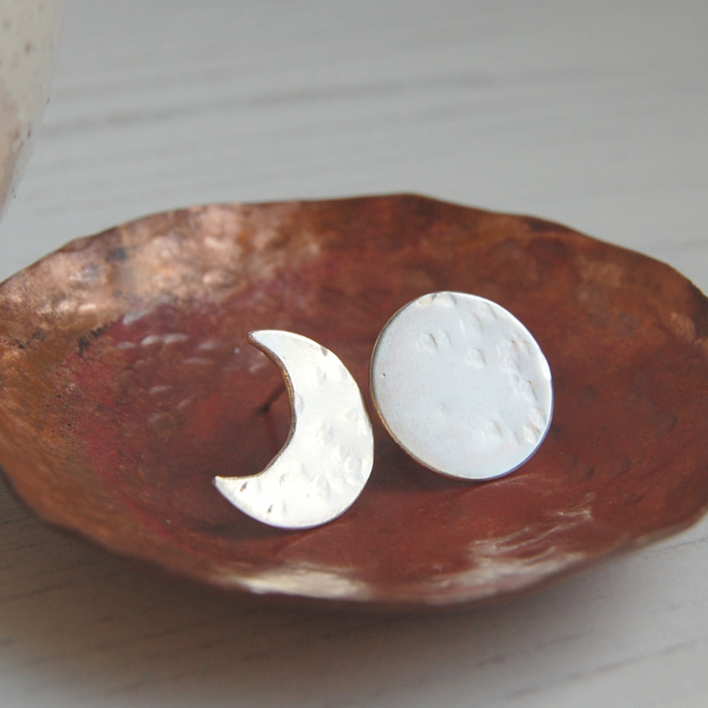 A pair or silver stud earrings, one a full moon with some hammered marks and one a crescent moon with some hammered marks sat in a copper dish, by zoe ruth designs.