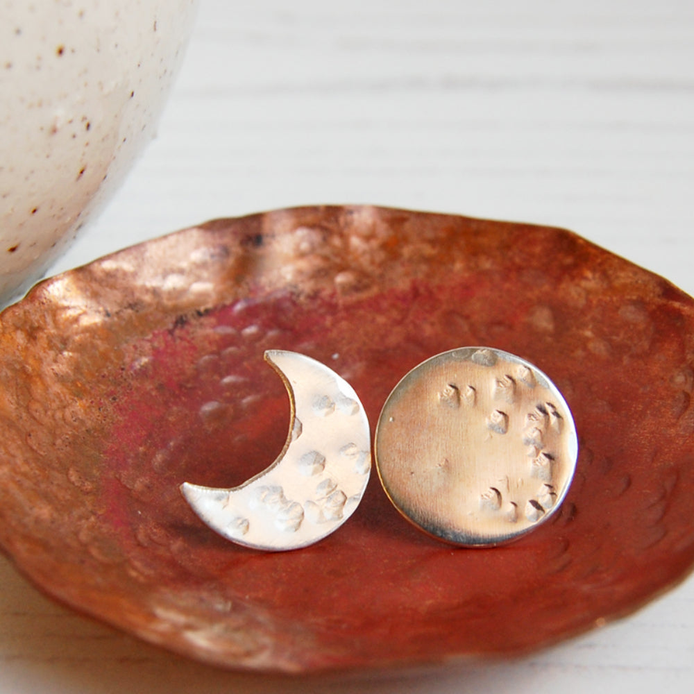A pair or silver stud earrings sat in a copper dish, one a full moon with some hammered marks and one a crescent moon with some hammered marks, by zoe ruth designs.