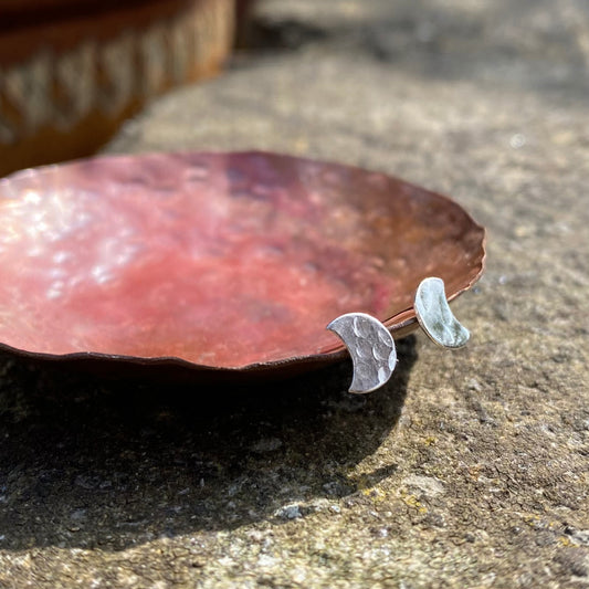 A pair of small silver hammered stud earrings in the shape of a crescent moon, resting on a copper dish, by zoe ruth designs.