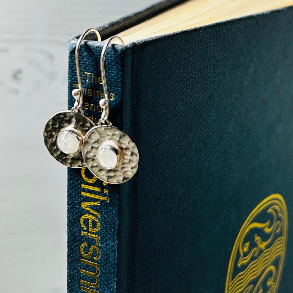 A pair of handmade silver earring hooks from which hangs a hammered flat silver disk and a round facet cut moonstone in the centre, by zoe ruth designs