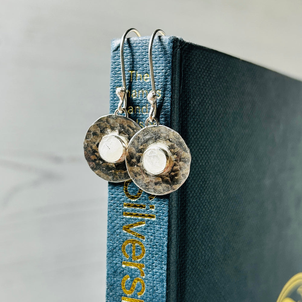 A pair of handmade silver earring hooks from which hangs a hammered flat silver disk and a round facet cut moonstone in the centre, displayed hanging on a book spine by zoe ruth designs