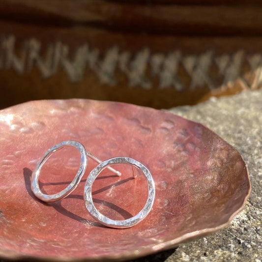 Showing a pair of stud earrings in silver wire formed into a circle outline and hammered to catch the light, sitting in a copper dish, by zoe ruth designs.