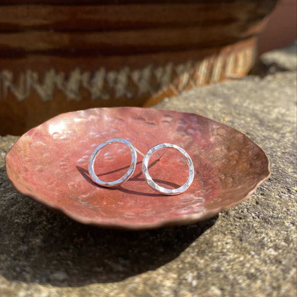 Showing a pair of stud earrings in silver wire formed into a circle outline and hammered to catch the light, sitting in a copper dish, by zoe ruth designs.