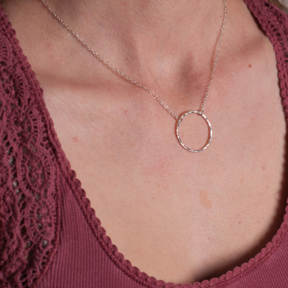 A slim circle frame pendant, sterling silver and lightly hammered, hangs from a sterling silver chain, shown being worn by zoe ruth designs.