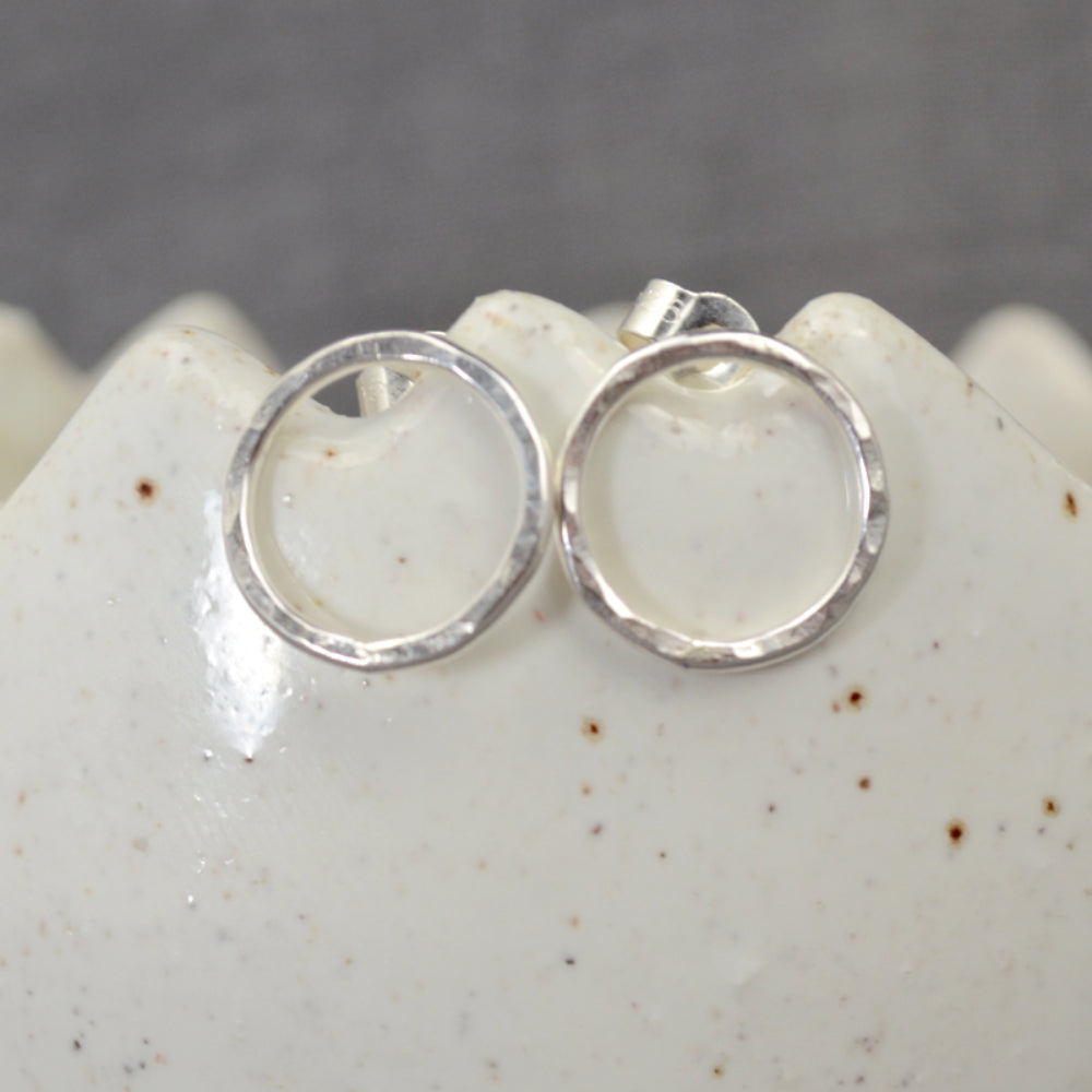 Showing a pair of stud earrings in silver wire formed into a circle outline and hammered to catch the light, sat on the edge of a ceramic dish, by zoe ruth designs.