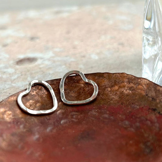 A pair of silver tud earrings in the shape of the outline of a heart, hammered surface, resting on a copper dish, by zoe ruth designs.