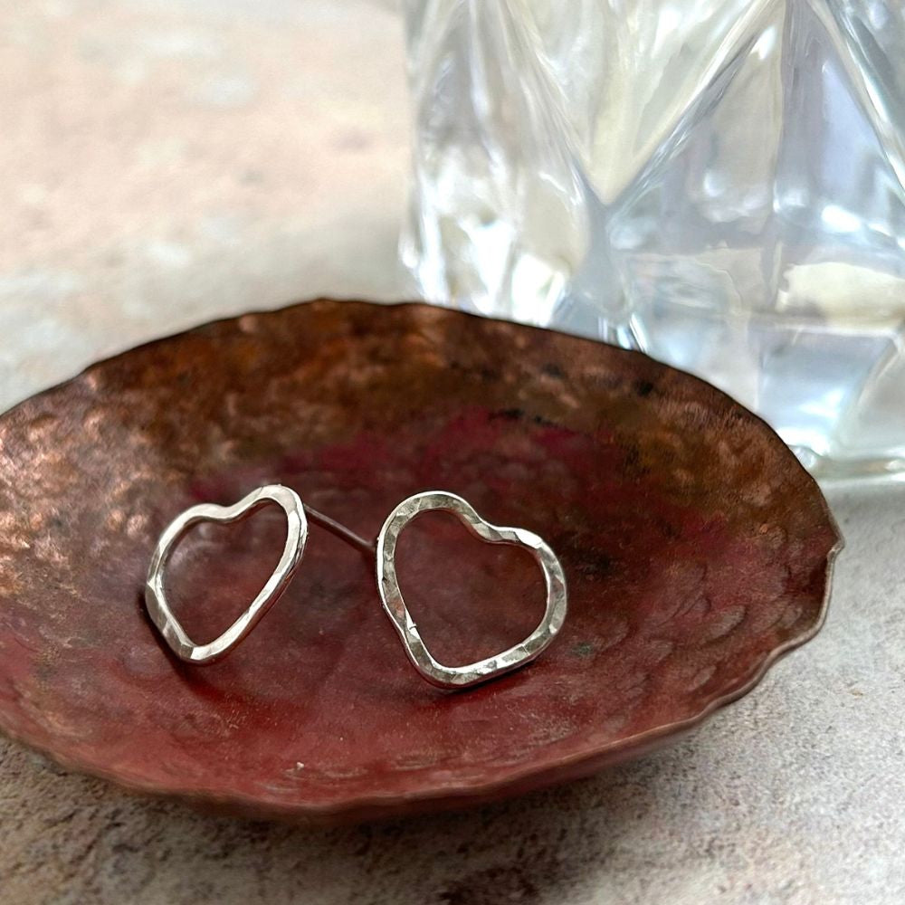 A pair of silver tud earrings in the shape of the outline of a heart, hammered surface, resting on a copper dish, by zoe ruth designs.