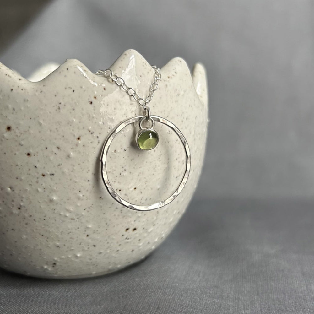 A 5mm fresh green Peridot stone set in silver hangs loosely at the top of a larger slim and hammered circle frame as the pendant of this necklace by zoe ruth designs.