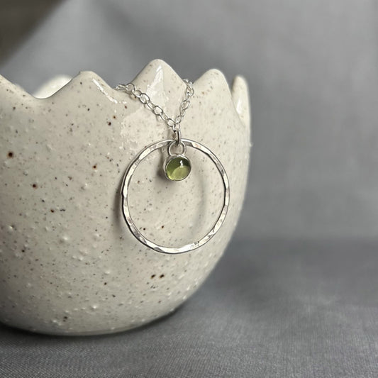 A 5mm fresh green Peridot stone set in silver hangs loosely at the top of a larger slim and hammered circle frame as the pendant of this necklace by zoe ruth designs.