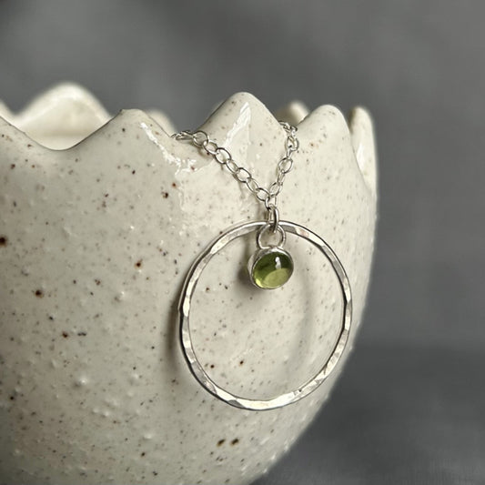 A 5mm fresh green Peridot stone set in silver hangs loosely at the top of a larger slim and hammered circle frame as the pendant of this necklace by zoe ruth designs.