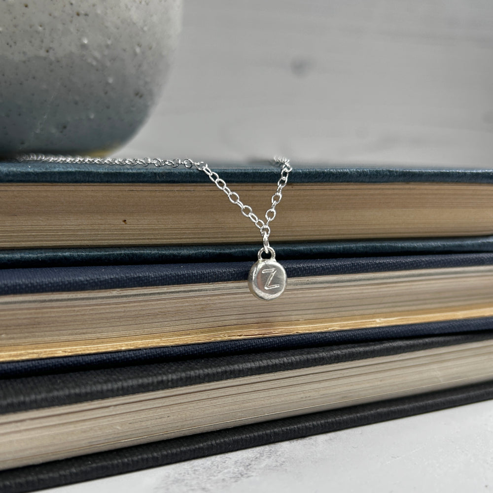 Showing a single silver handmade pebble with a stamped letter on it, hanging as a charm from a silver chain, by zoe ruth designs