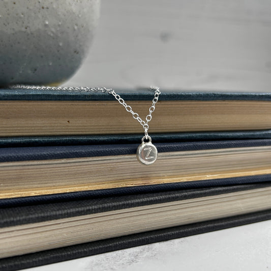 Showing a single silver handmade pebble with a stamped letter on it, hanging as a charm from a silver chain, by zoe ruth designs