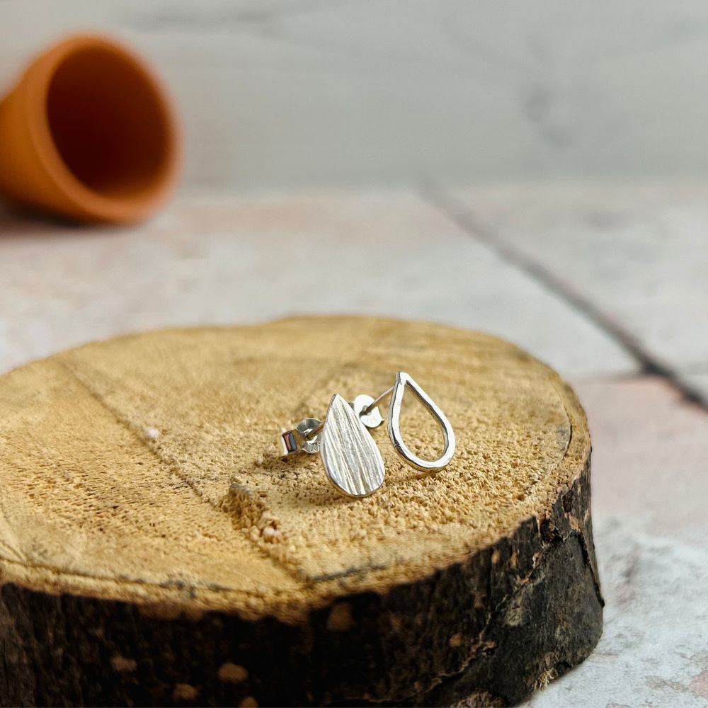 Handmade small silver stud earrings, one a solid teardrop shape with vertical lines texture, the second the outline fo a teardrop shape with dimple hammered texture. Displayed sat on sawn log with a terracotta plantpot in the background