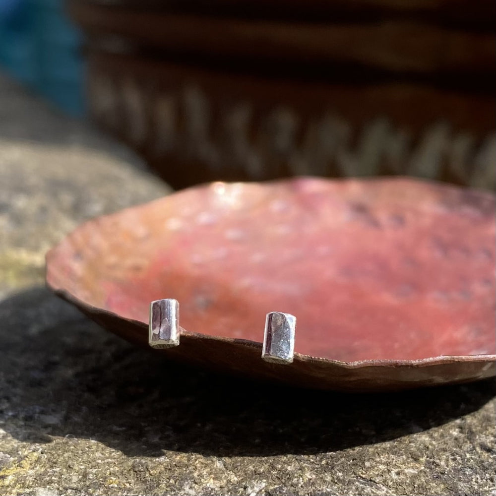 A pair of small silver hammered rectagle stud earrings, shown resting on a copper dish in the sunshine, by zoe ruth designs.