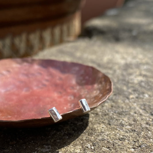 A pair of small silver hammered rectagle stud earrings, shown resting on a copper dish in the sunshine, by zoe ruth designs.
