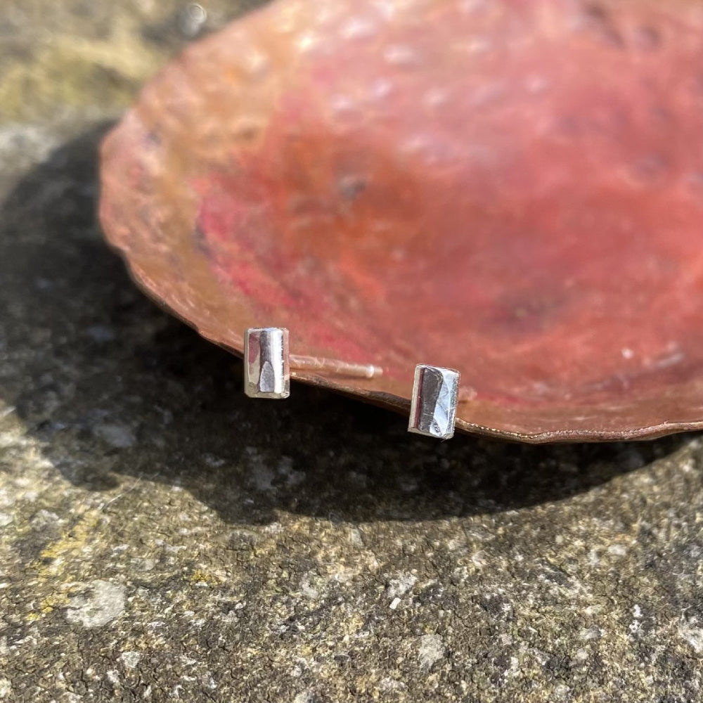 A pair of small silver hammered rectagle stud earrings, shown resting on a copper dish in the sunshine, by zoe ruth designs.