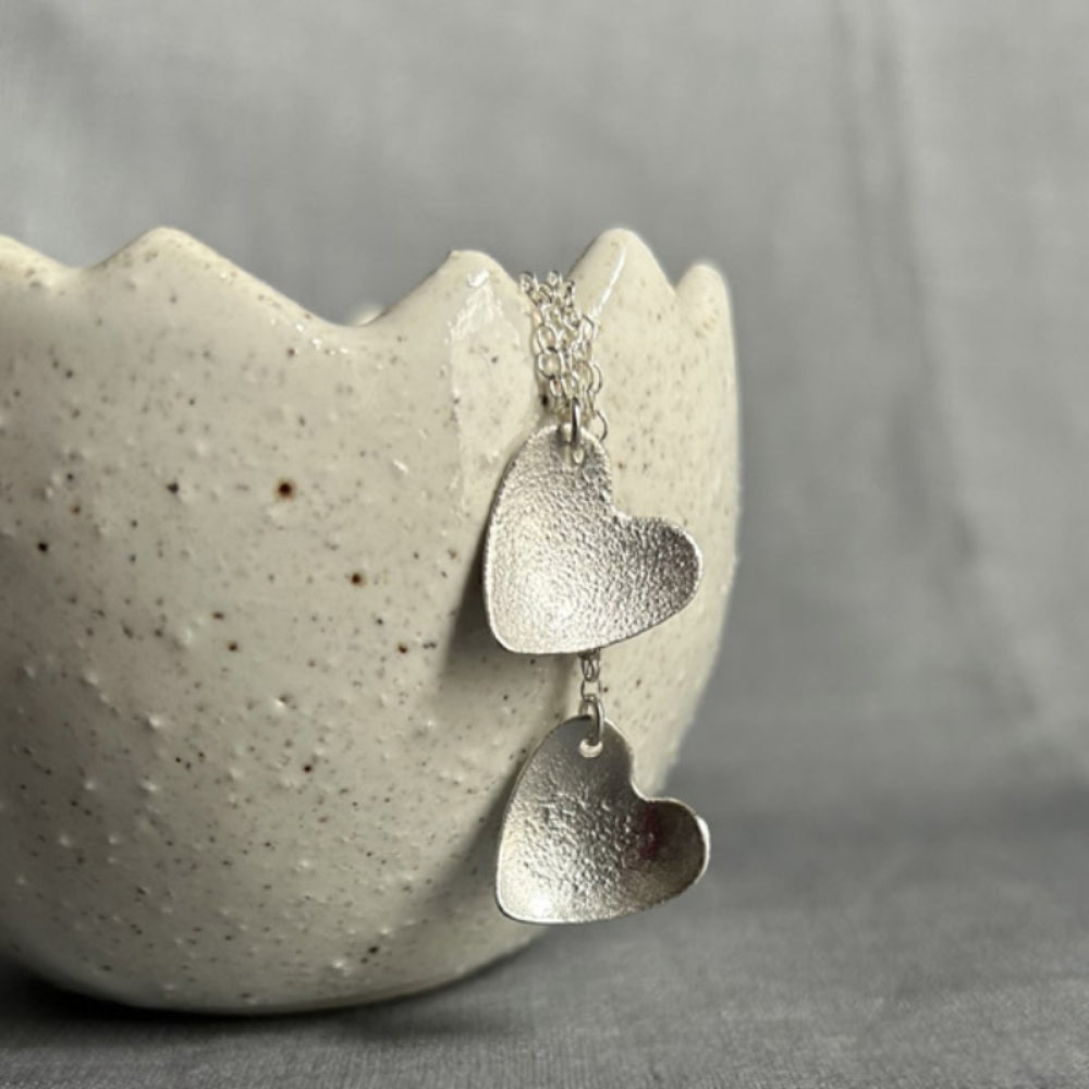 Two pendants with a bumpy surface textured silver heart hanging from a silver chains, showing the difference in textures, displayed hangng from a ceramic pot by zoe ruth designs.