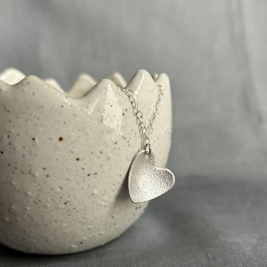 A bumpy surface textured silver heart hangs as a pendant from a silver chain, displayed hanging from a ceramic pot by zoe ruth designs.